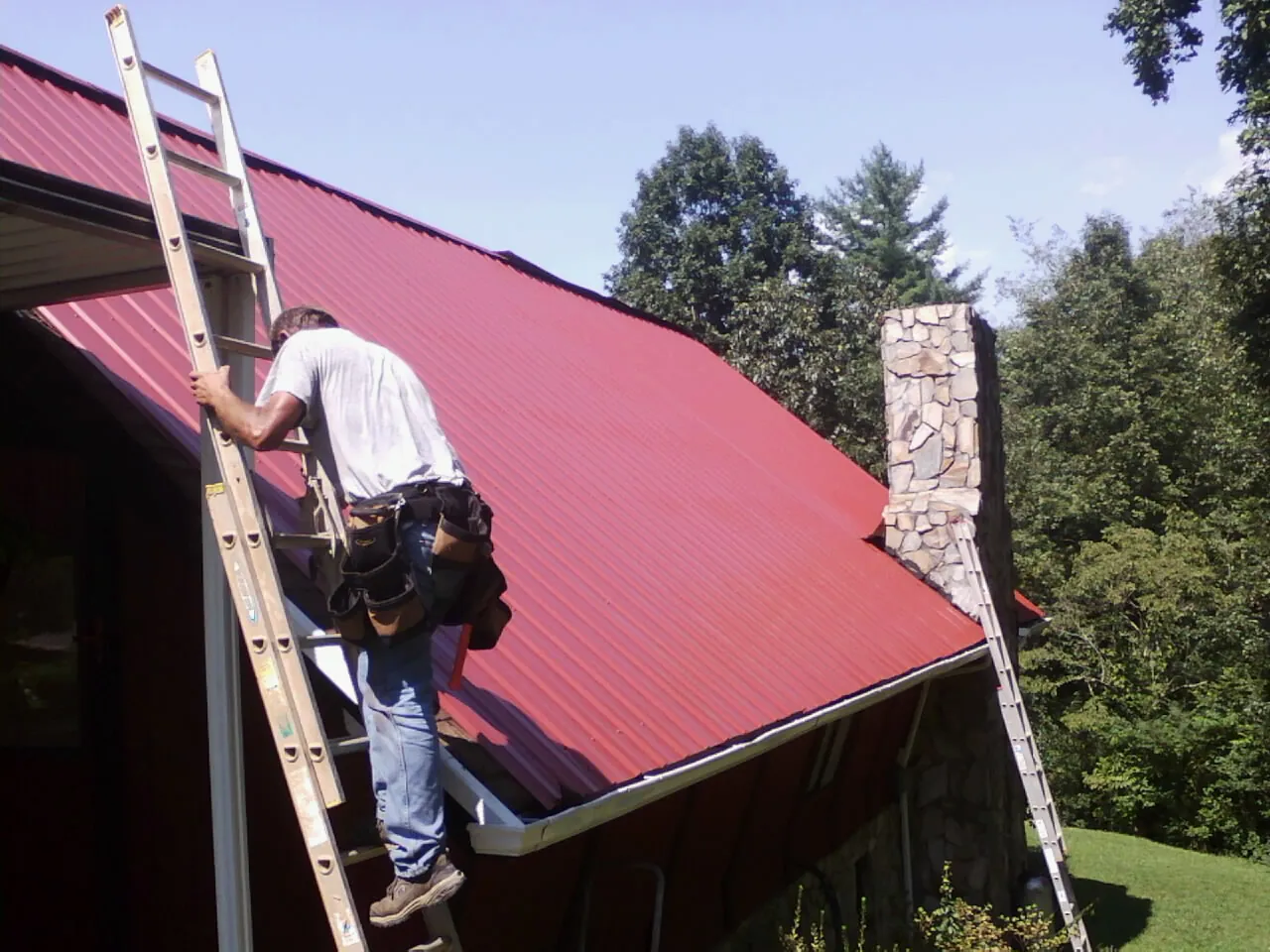 Roofer on a ladder beside a red metal roof and stone chimney — Soffit & Fascia Repair by Roofing Brothers in Higginsville